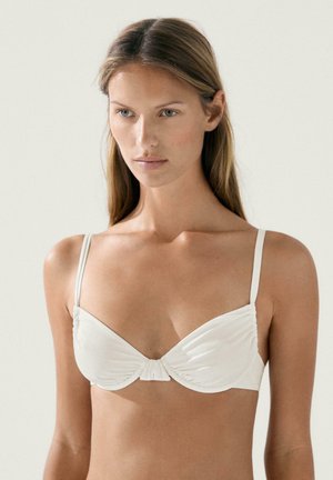 Žena s dlouhými blond vlasy, která nosí bílou podprsenku s tenkými ramínky, čelem k nám na jednobarevném světlém pozadí.