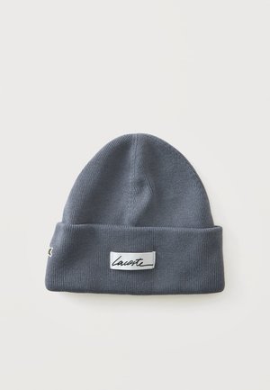 Gray knitted beanie hat with folded cuff and white rectangular label showing black cursive "Lacoste" text on front.