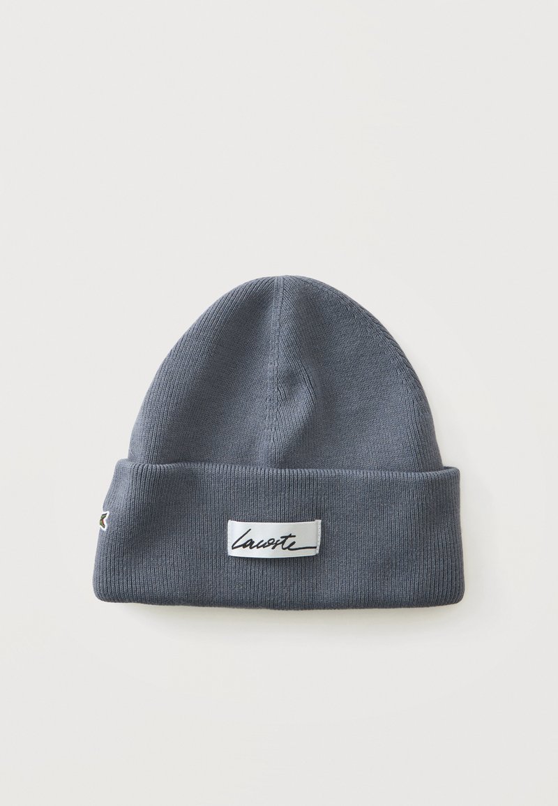 Bonnet en maille gris de type beanie avec revers plié et étiquette rectangulaire blanche affichant un texte en cursive noir « "Lacoste" » à l’avant.