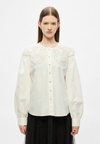 Biela bavlnená košeľa s dekoratívnym vyšívaným golierom, dlhými nafúknutými rukávmi a zapínaním na gombíky vpredu. Má detailné čipkové doplnky a rovný lem.