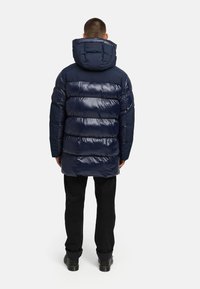 Laivastonsininen puffatakki kiiltävällä pinnalla, jossa on huppu ja vaakasuuntainen tikkausmuotoilu. Yhdistetty mustiin housuihin ja kestävään kenkiin.