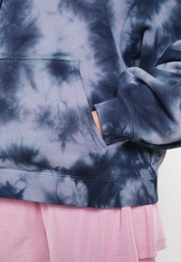 Felpa tie-dye blu navy e celeste realizzata in tessuto morbido, con tasca a marsupio e polsini a coste, abbinata a un indumento rosa sotto.