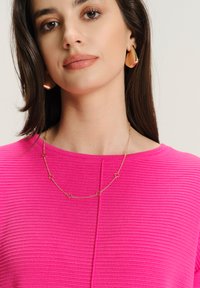 Młoda kobieta z prostymi brązowymi włosami nosi teksturowany, jasnoróżowy top, złotą geometryczną naszyjnik i masywne złote kolczyki w kształcie kropli.