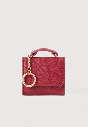 Bolsa de couro texturizado vermelho com uma alça curta e um acessório em corrente dourada. Apresenta um detalhe de anel metálico circular. Forma compacta e quadrada.