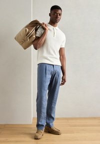 Cămașă polo crem cu dungi, pantaloni din in albaștri deschiși și încălțăminte bej. Poartă o geantă de voiaj bej cu accente maronii. Fundal din podea din lemn.