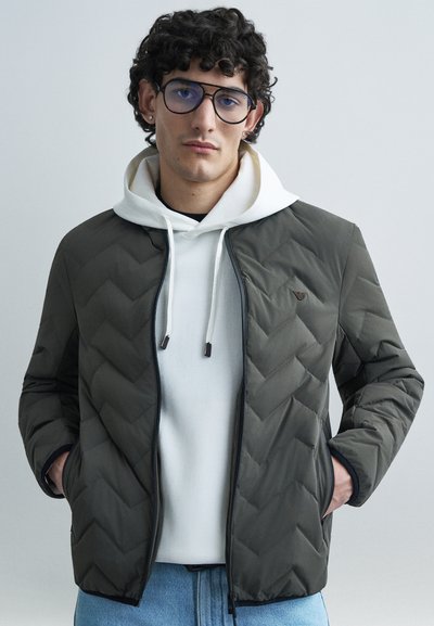 Donkergroene gewatteerde jas met visgraatpatroon, zwarte ritssluiting en twee zijzakken, gedragen over een witte hoodie met trekkoorden.