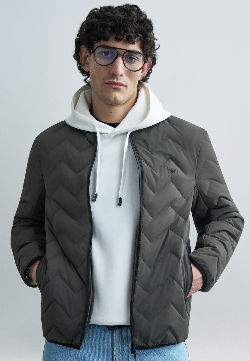 Tmavozelená prešívaná bunda so vzorom chevron, čiernym zipsom a dvoma bočnými vreckami, cez bielu mikinu s kapucňou a sťahovacími šnúrkami.