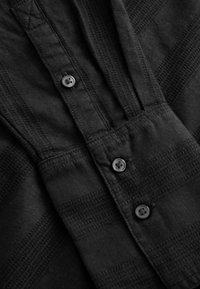 Chemise noire avec un motif texturé, dotée d'une patte de boutonnage, de quatre boutons noirs et de détails de coutures visibles. Tissu lisse.