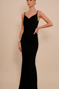 Femeie îmbrăcată într-o rochie de seară neagră, ajustată, fără mâneci, cu bretele subțiri, brățară de aur și cercei, pozând pe un fundal bej.