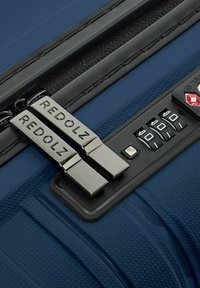 Doppelte silberne Reißverschluss-Zipper mit der Aufschrift "REDOLZ" auf einem blauen strukturierten Koffer mit einem schwarzen dreistelligen Zahlenschloss.
