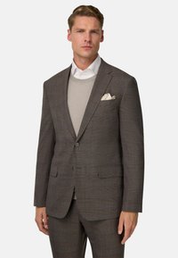 Completo a quadri marrone con pantaloni abbinati, rever a punta, chiusura con un bottone e fazzoletto da taschino di colore crema. Maglione grigio chiaro sotto.
