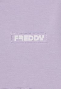 Parche con el logotipo de Freddy sobre un fondo de tela de color púrpura claro. El parche muestra texto blanco en una etiqueta rectangular cosida. Textura suave.