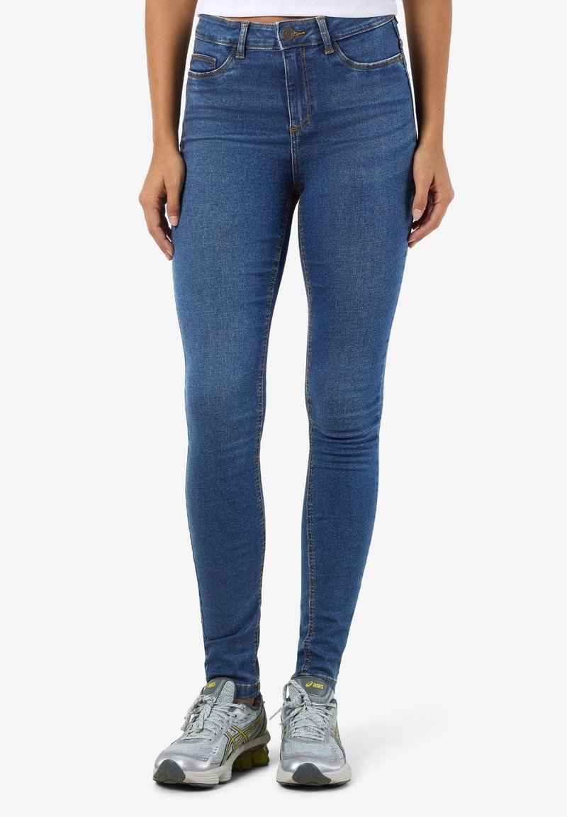 Femme portant un jean skinny bleu et des chaussures de course argentées, debout avec les mains détendues le long du corps sur un fond blanc.