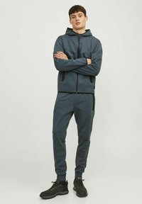 Mørkegrå zip-up hættejakke med hætte og sidelommer, parret med matchende tapered joggers. Begge har et glat, tekstureret stof og kontrasterende sorte detaljer.