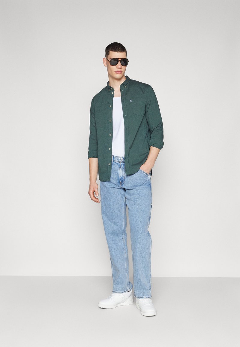 Camisa verde de botones sobre una camiseta blanca, vaqueros de corte relajado de color azul claro, y zapatillas blancas; lleva gafas de sol negras; fondo liso.