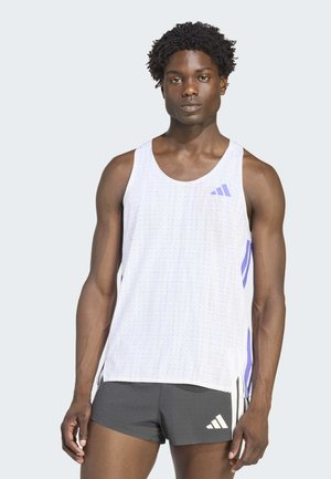 Atletyczny mężczyzna z krótkimi kręconymi włosami ubrany w biały sportowy tank top bez rękawów oraz czarne krótkie spodenki sportowe z logo marki na jasnym tle.