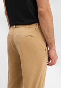 Beżowe spodnie sztruksowe z pionowymi prążkami, wyposażone w tylne kieszenie i teksturowaną powierzchnię, zestawione z czarną koszulą.