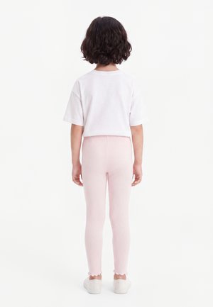 Barn iført en hvid kortærmet skjorte, lyserøde leggings med flæsekant og hvide sneakers, stående med ryggen til på en hvid baggrund.