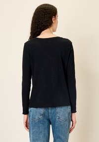 Ženska s kodrastimi lasmi nosi preprosto črno majico z dolgimi rokavi in modre denim jeans hlače, stoji s hrbtom obrnjena proti kameri na svetlo bež ozadju.