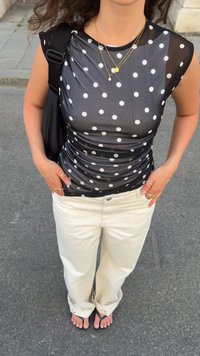Svart genomskinlig topp med vita prickar, rynkad design; kombinerad med krämfärgade vida byxor och flip-flop-sandaler. Tillbehören inkluderar lager på lager halsband.