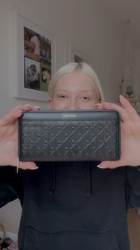 Čierna kožená peňaženka s textúrovaným vzorom prepletených písmen, so zipsovým zatváraním a kovovým logom na prednej strane.