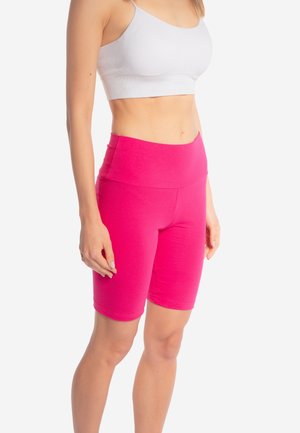 Kobieta w białym krótkim topie i jasnoróżowych, wysokim stanem krótkich spodenkach rowerowych, stojąca na tle jednolitego białego tła.