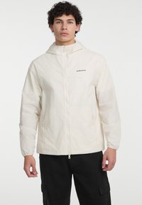 Jachetă ușoară bej cu glugă, fermoar vizibil și procese elastice la mansete. Are logo pe piept. Purtată peste pantaloni cargo negri.