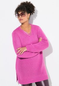 Pull rose en maille douce avec un col en V, coupe oversize, poignets et ourlet côtelés, présentant des fentes latérales pour plus de liberté de mouvement ; texture superposée au niveau du col.