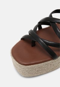 Sandale à plateforme en cuir noir avec des lanières entrecroisées, semelle intérieure marron et semelle en jute tissée, présentant une finition texturée et un design plat.