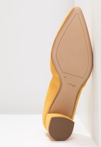 Chaussure jaune en daim à bout pointu, inclinée pour montrer la semelle lisse couleur fauve avec un petit talon et le nom de la marque « högl » embossé près de la voûte.