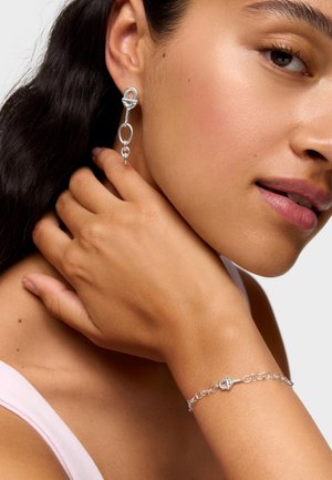 Donna che indossa un orecchino a catena d'argento e un braccialetto abbinato, mano posata sul collo, trucco delicato, capelli lunghi e scuri, spallina di top rosa chiaro visibile.