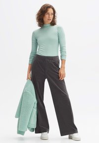 Licht blauwgroene lange mouwen top met ribbeltextuur, gecombineerd met donkergrijze wijde corduroy broek. Model houdt een bijpassende blauwgroene fleecejas vast.