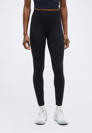 Άτομο που φοράει μαύρα ψηλόμεσα κολάν, λευκές κάλτσες και λευκά αθλητικά παπούτσια, στέκεται μπροστά σε ένα απλό φωτεινό φόντο.