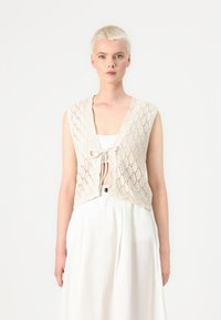 Knit, sleeveless cream vest with diamond pattern and tie closure at the front, paired with a plain white dress.