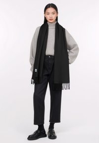 Sciarpa nera con dettagli frangiati indossata sopra un dolcevita grigio chiaro e jeans scuri. La sciarpa è realizzata in un materiale morbido che offre calore.