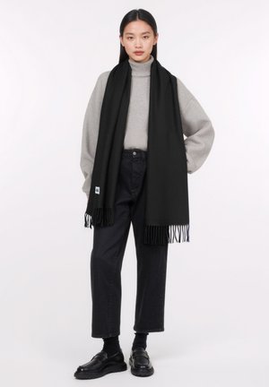 Zwarte sjaal met franjes, gedragen over een lichtgrijze turtleneck en donkere jeans. De sjaal is gemaakt van een zacht materiaal en biedt warmte.