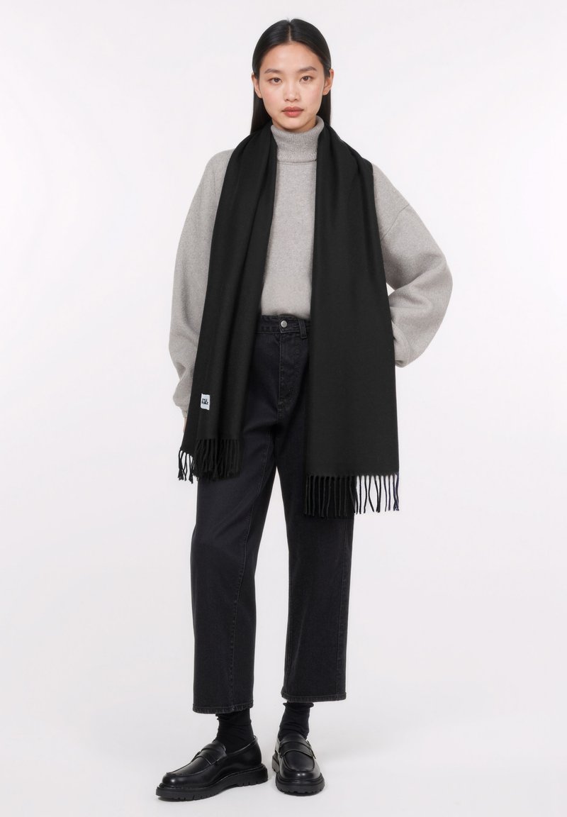 Sciarpa nera con dettagli frangiati indossata sopra un dolcevita grigio chiaro e jeans scuri. La sciarpa è realizzata in un materiale morbido che offre calore.