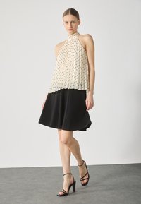 Cream pleated halter top with black polka dots, paired with a black skater skirt. Model wears strappy black heels. Simple background.