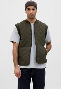 Quiltet olivengrøn ærmeløs vest med lynlås foran, der har to sidelommer og en rund halsudskæring, båret over en grå T-shirt.
