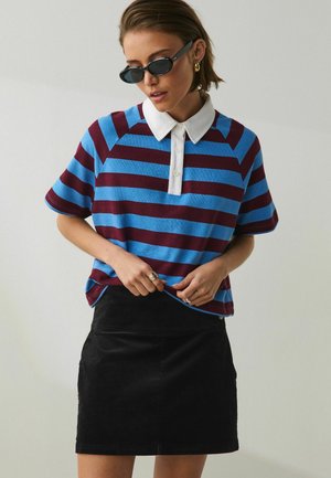 Frau mit schwarzen Sonnenbrillen, blau und bordeaux gestreiftem Hemd mit weißem Kragen und schwarzem Rock vor einem schlichten hellen Hintergrund.