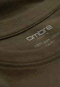 Olivově zelená bavlněná tkanina s hladkou strukturou, s kulatým výstřihem a tištěnou štítkem s péčí, na kterém je bíle napsáno "100 % čistá bavlna".