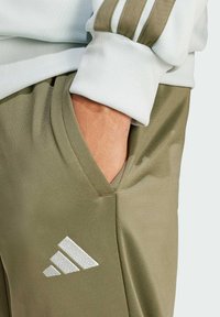 Olivgröna byxor med en slät textur, med en sidficka och vit Adidas-logotyp. Åtföljd av en ljusblå, strukturerad topp.