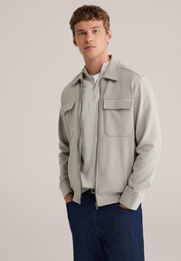 Hall hall lukuga jakk kahe ees taskuga, kantud helehallil poole luku kampsunil, kombineeritud tumesiniste teksapükstega.