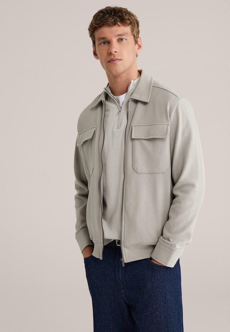 Hall hall lukuga jakk kahe ees taskuga, kantud helehallil poole luku kampsunil, kombineeritud tumesiniste teksapükstega.