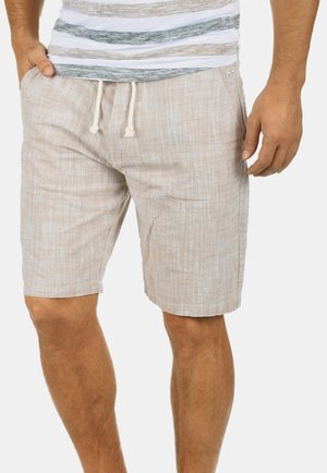 Man draagt beige linnen shorts met trekkoord en een wit T-shirt met grijze en bruine horizontale strepen.