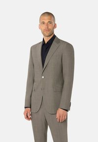 Mees lühikeste juustega, hallikaspruuni ülikonna ja musta särgiga, seismas lihtsa heledahalli tausta ees.