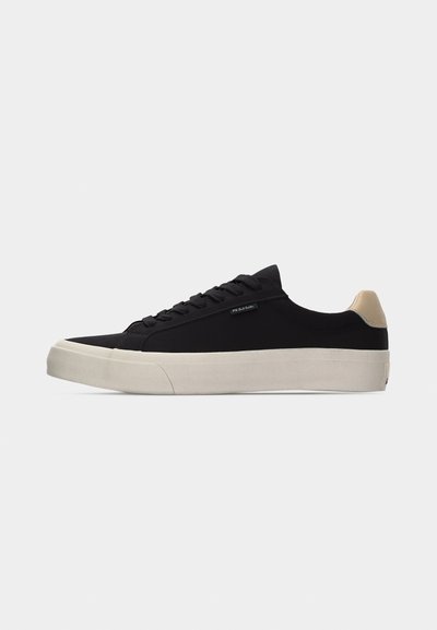 Μαύρο sneaker χαμηλό με μπεζ ταμπελάκι στη φτέρνα, λευκή παχιά ελαστική σόλα, μαύρα κορδόνια και μικρή ετικέτα "PS Paul Smith" στο πλάι.