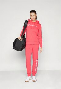 Raudonas džemperis su kapišonu ir laisvalaikio kelnės su baltu "Champion" logotipu, derinama su juoda sportine krepšiu ir baltais sportbačiais. Lygus audinys ir laisvas kirpimas.