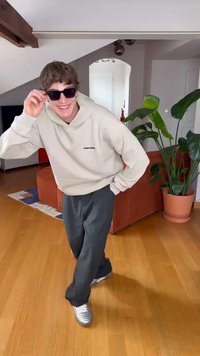 Pelēks kapuci bezrāvējs ar logotipu, pelēki bikses un sudraba sporta apavi; valkājot melnas saulesbrilles; iekštelpu vide ar augiem un mēbelēm.