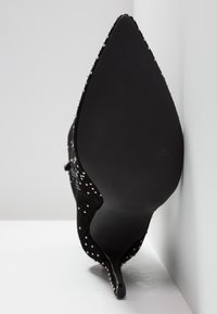 Chaussure à talon haut en daim noir avec un bout pointu, ornée de clous argentés, dotée d'une semelle texturée. La vue est centrée sur le talon et la semelle.